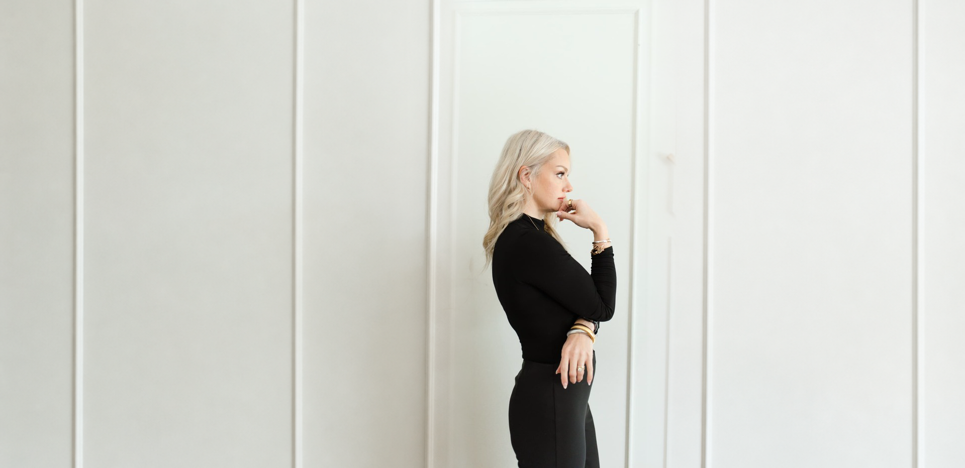 April Martini standing in profile with her hand at her chin, in front of a white paneled wall and staircase